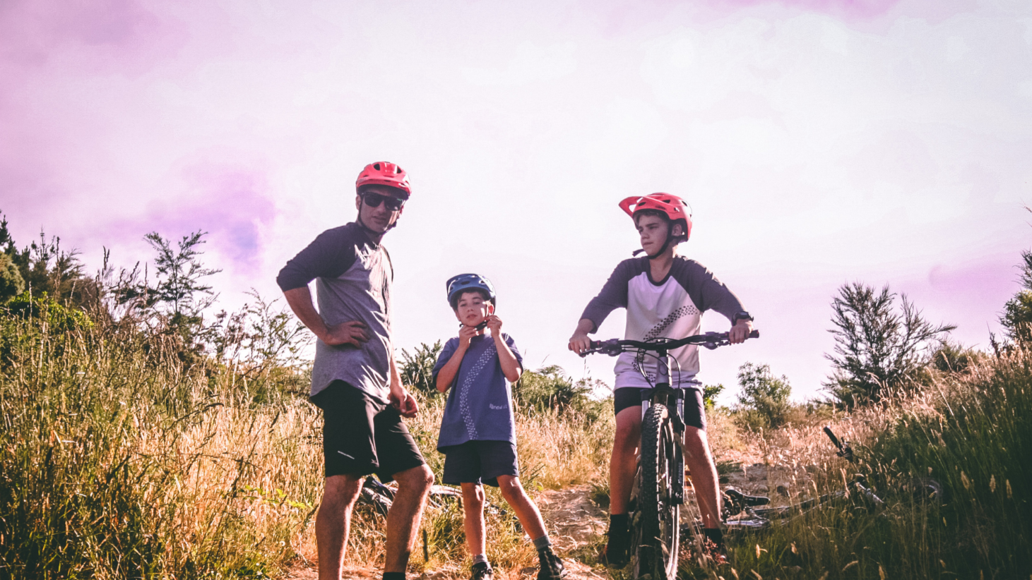 Three cyclists wearing helmets ride through tall grass on a sunny day, with one child on a bicycle i