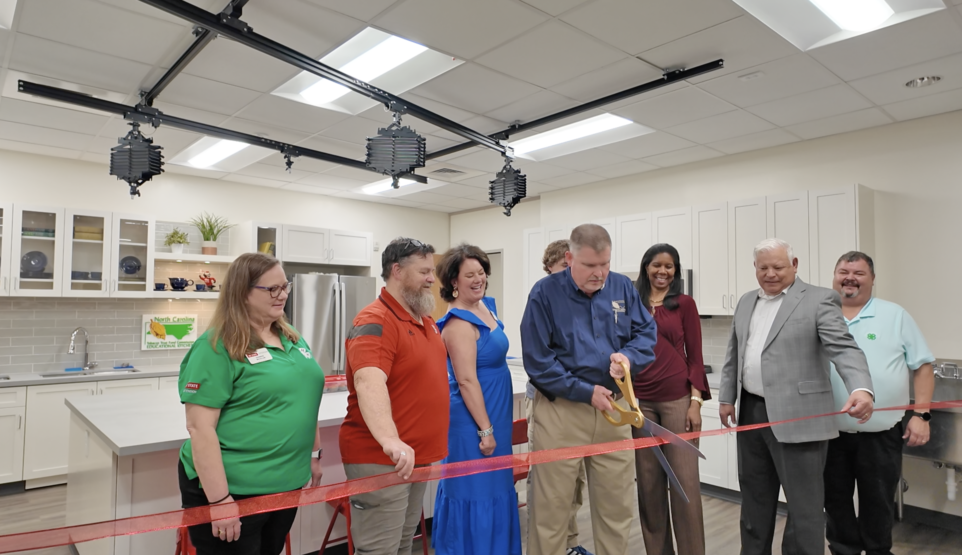 8 individuals cutting a ribbon in a kitchen